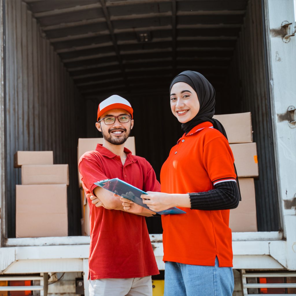 Two movers in Dubai wearing red uniforms standing near a truck, smiling and holding a clipboard.